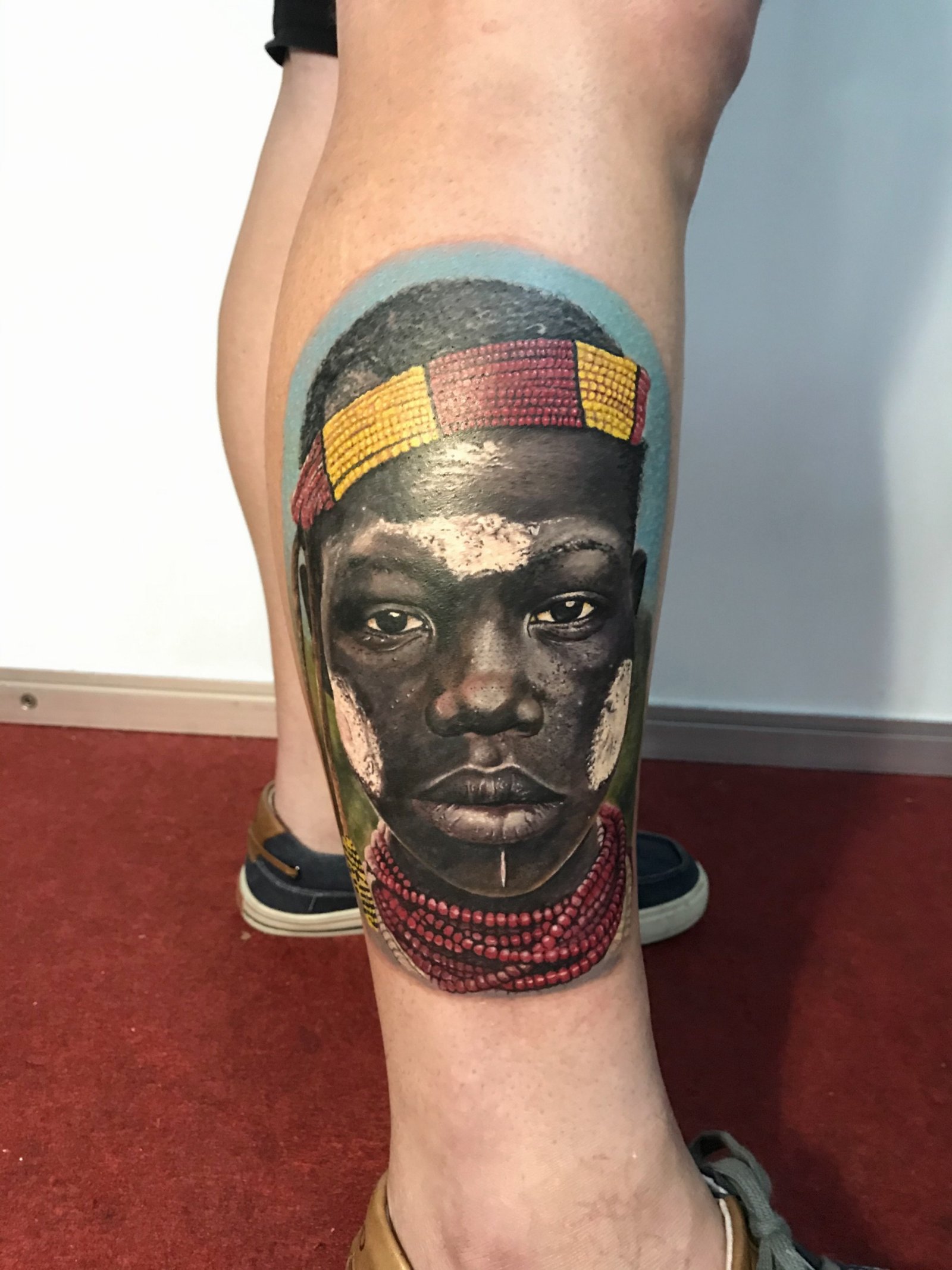 10wynj26xss1yzvpr6ixwma Tatuagem realista de rosto com detalhes coloridos na panturrilha de uma pessoa em pé sobre carpete vermelho.