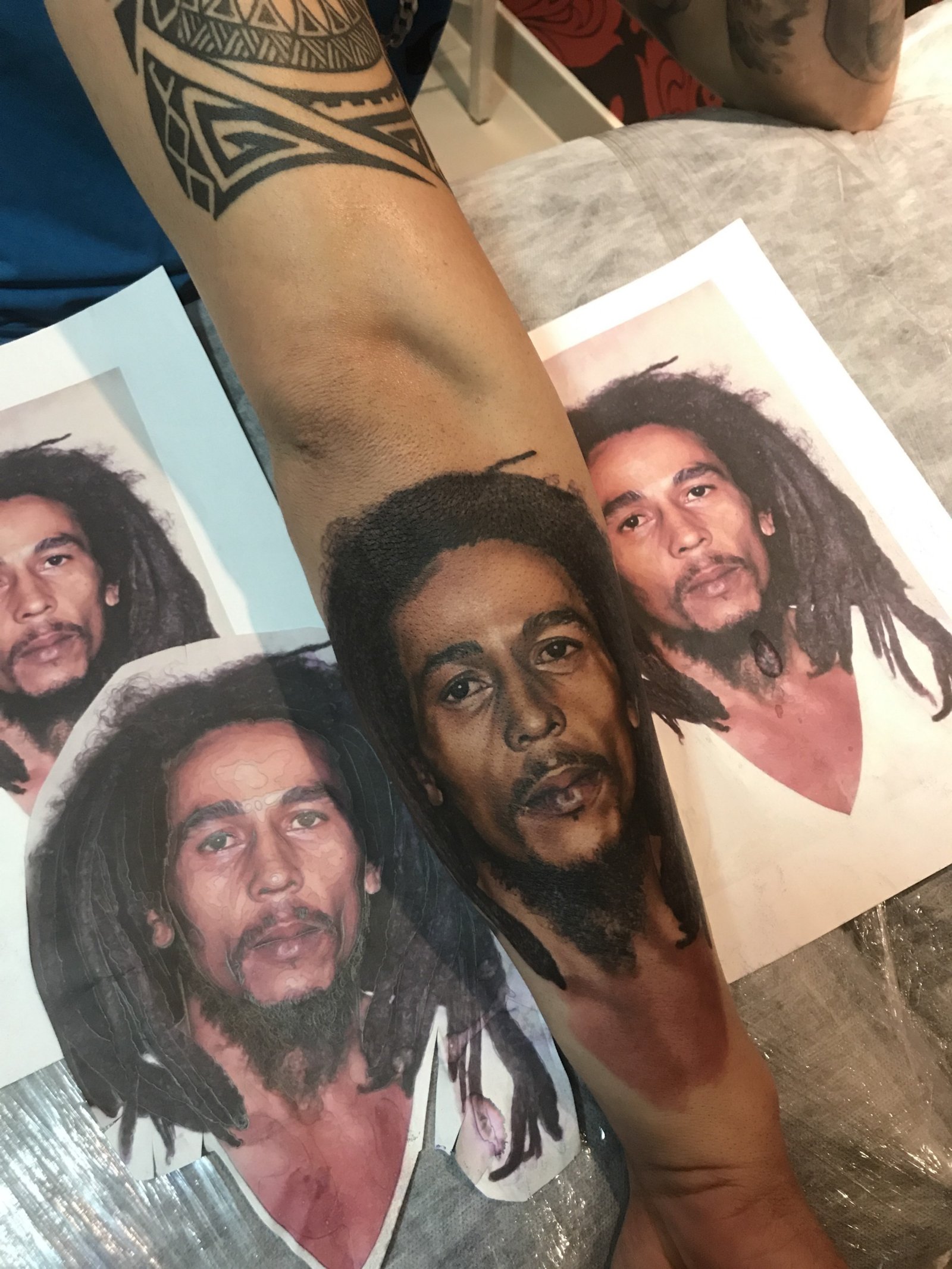 162ryjd0w0iy4q_vlsiesrg-1 Braço com tatuagem realista de um rosto masculino, ao lado de fotos de referência impressas.