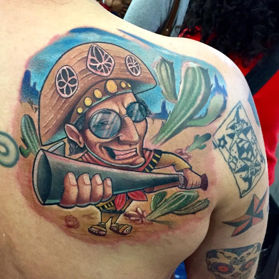 18r3dexqslayyt8o4scqexq-1 Uma tatuagem colorida de personagem caricatural com chapéu, óculos e megafone, em cenário com cactos e cartas.