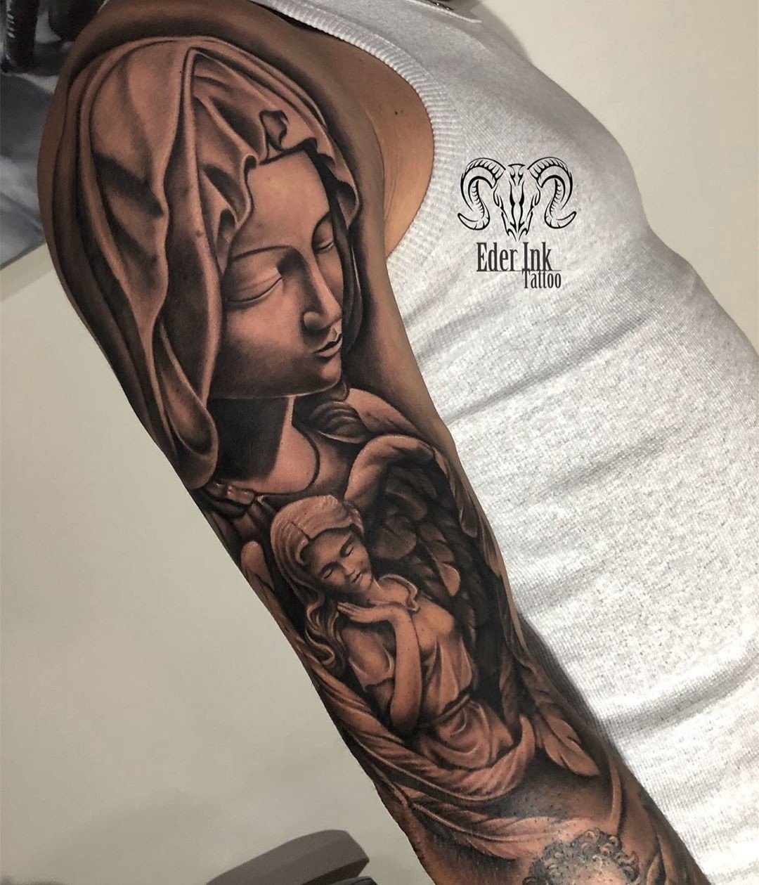 1_hnowvnmxgat6o8o4bcrng Desenho de tatuagem religiosa em braço, com figuras de anjo e pessoa com manto, em tons de preto e cinza.