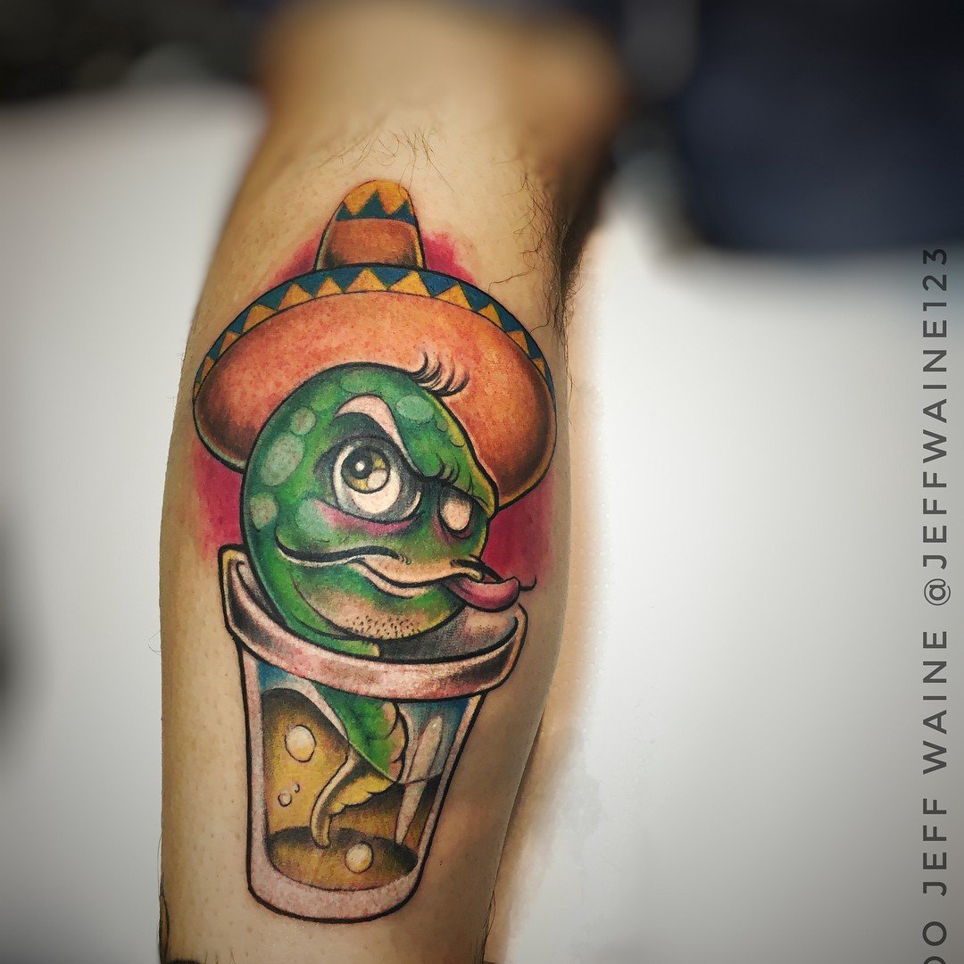 1c8huicgkl3g5m66nj7un8a-1 Tatuagem de uma criatura verde em um copo, usando um sombrero colorido, sobre uma perna.