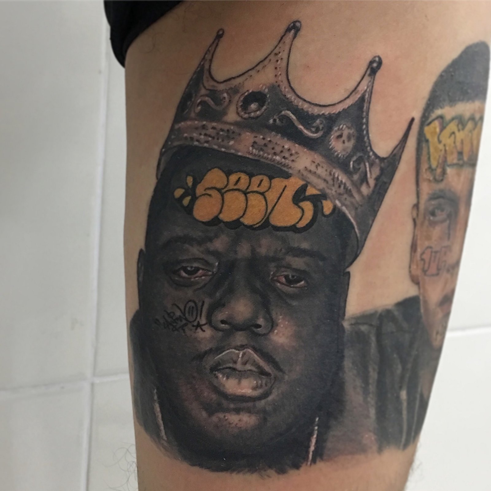 1caxhc3hnokchnftmli_c_g-1 Uma tatuagem de rosto masculino com coroa e inscrições, localizada na perna de uma pessoa.