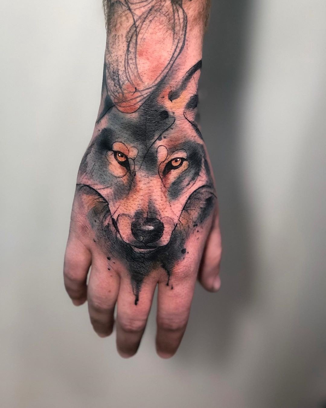 Mão com tatuagem de lobo em estilo aquarela, destacando olhos intensos e cores sombreadas.