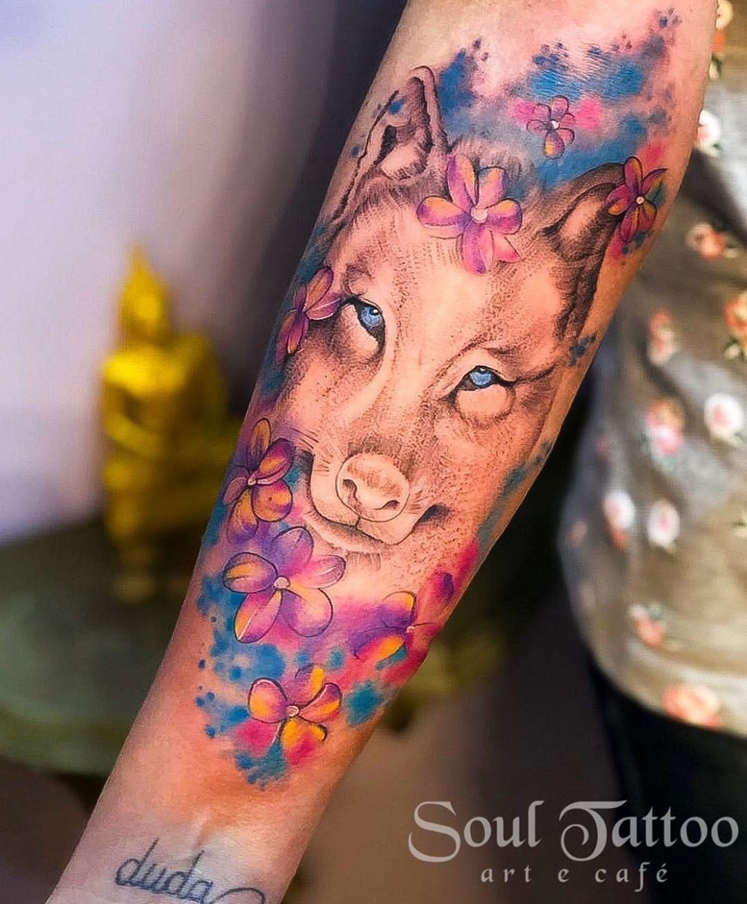 1ddc3irmedsi5em2g-dyjrw-1 Tatuagem colorida de lobo com flores roxas e azuis em um antebraço.