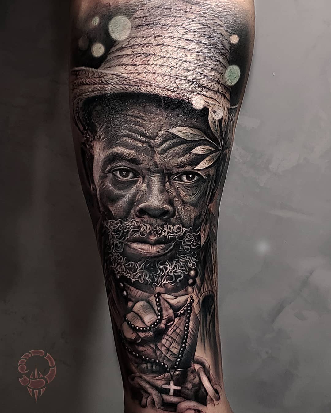 1duzcy_gt8whkb3_u1upy2w Um retrato detalhado de um rosto masculino com chapéu, adornado por folhas e colares, tatuado em um braço.