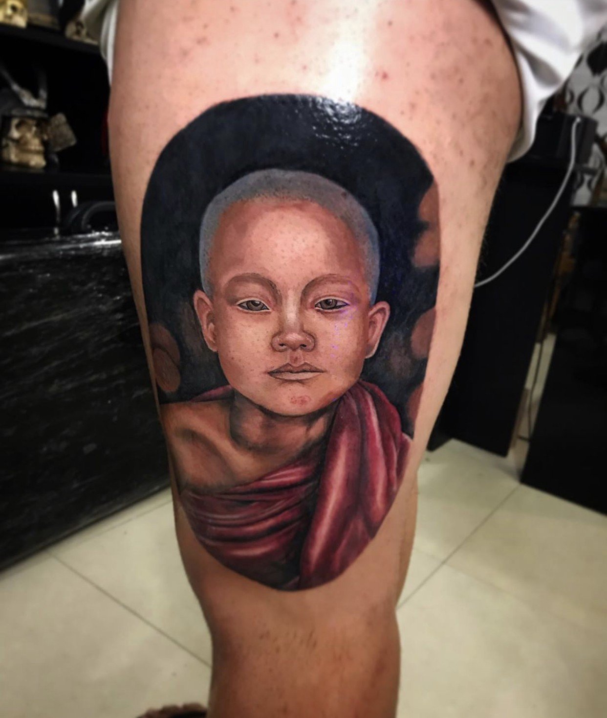 1e4cxayiyg0410gssdeycmg-1 Tatuagem realista de jovem monge em tons escuros, localizada na perna de uma pessoa.