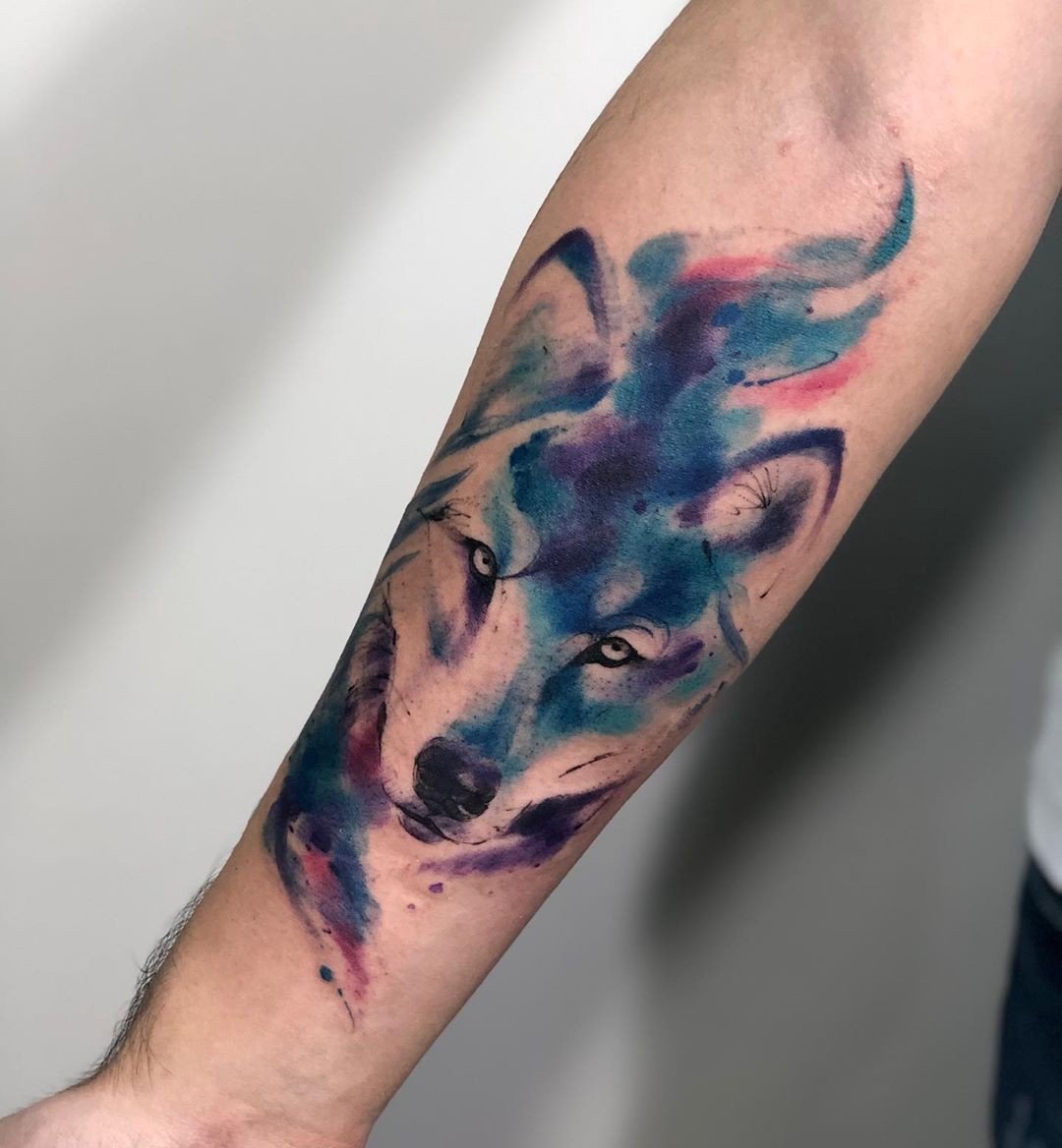 Braço com tatuagem de lobo em estilo aquarela, usando tons de azul, roxo e rosa.