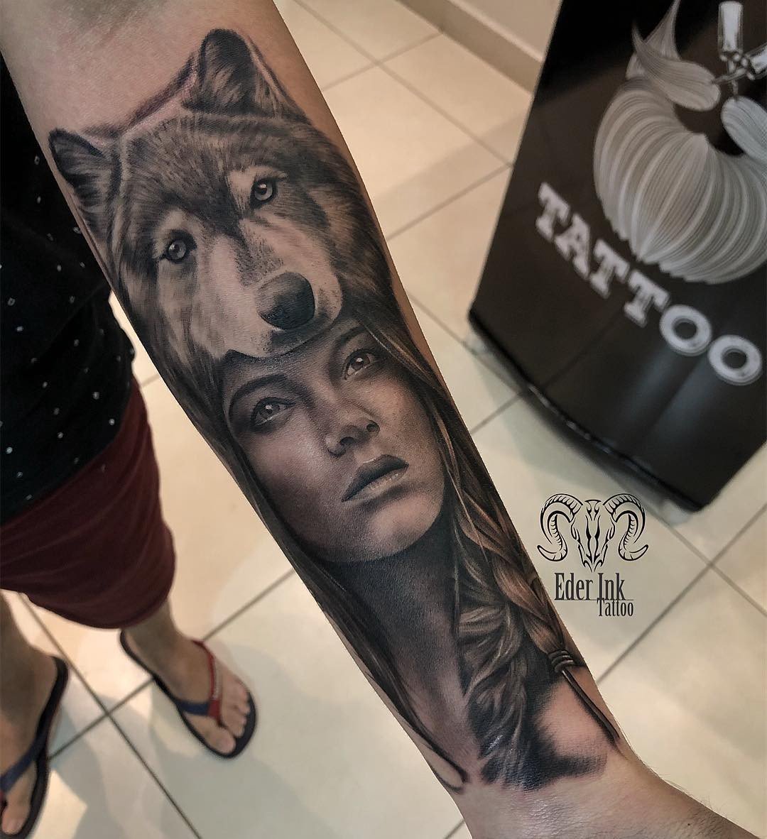 1er4o6iduv7deus6dr1o7cq Tatuagem realista de lobo e rosto feminino no antebraço de uma pessoa em pé sobre um piso claro.
