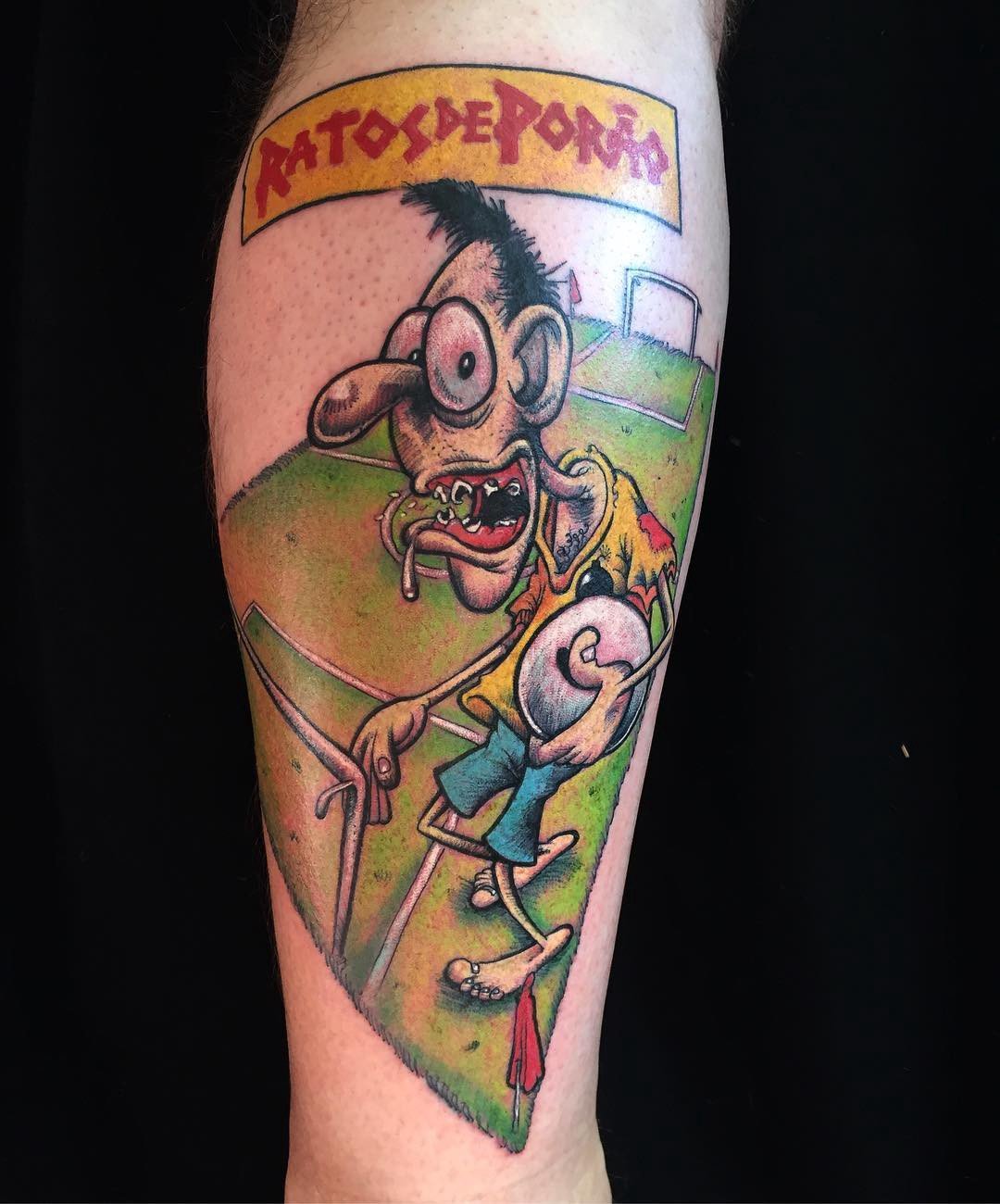 1ert6fi8zrzs5dlgcjnhu6a-1 Tatuagem colorida de personagem caricaturesco em campo de futebol, com texto estilizado acima.
