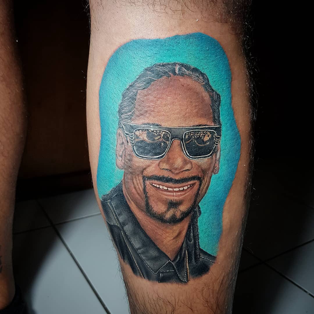 1fa2g9k8oppttrq1wyot4og-1 Desenho de rosto sorridente com óculos escuros, tatuado na perna, sobre fundo azul.