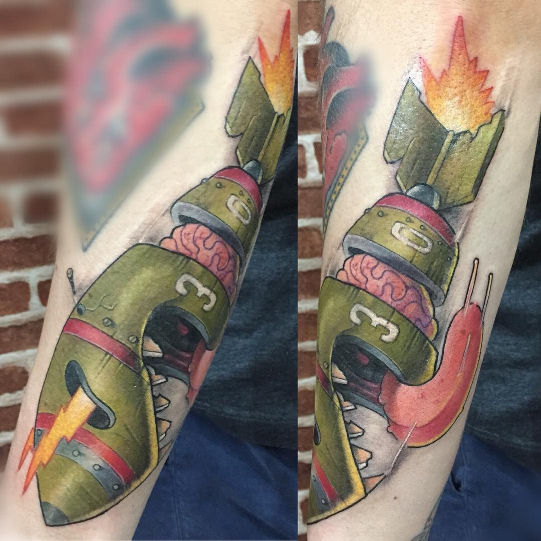 1gkhpskiyko74_5xe5vheza-1 Tatuagem colorida de robô com cérebro exposto e foguete, em braço, sobre fundo de tijolos.