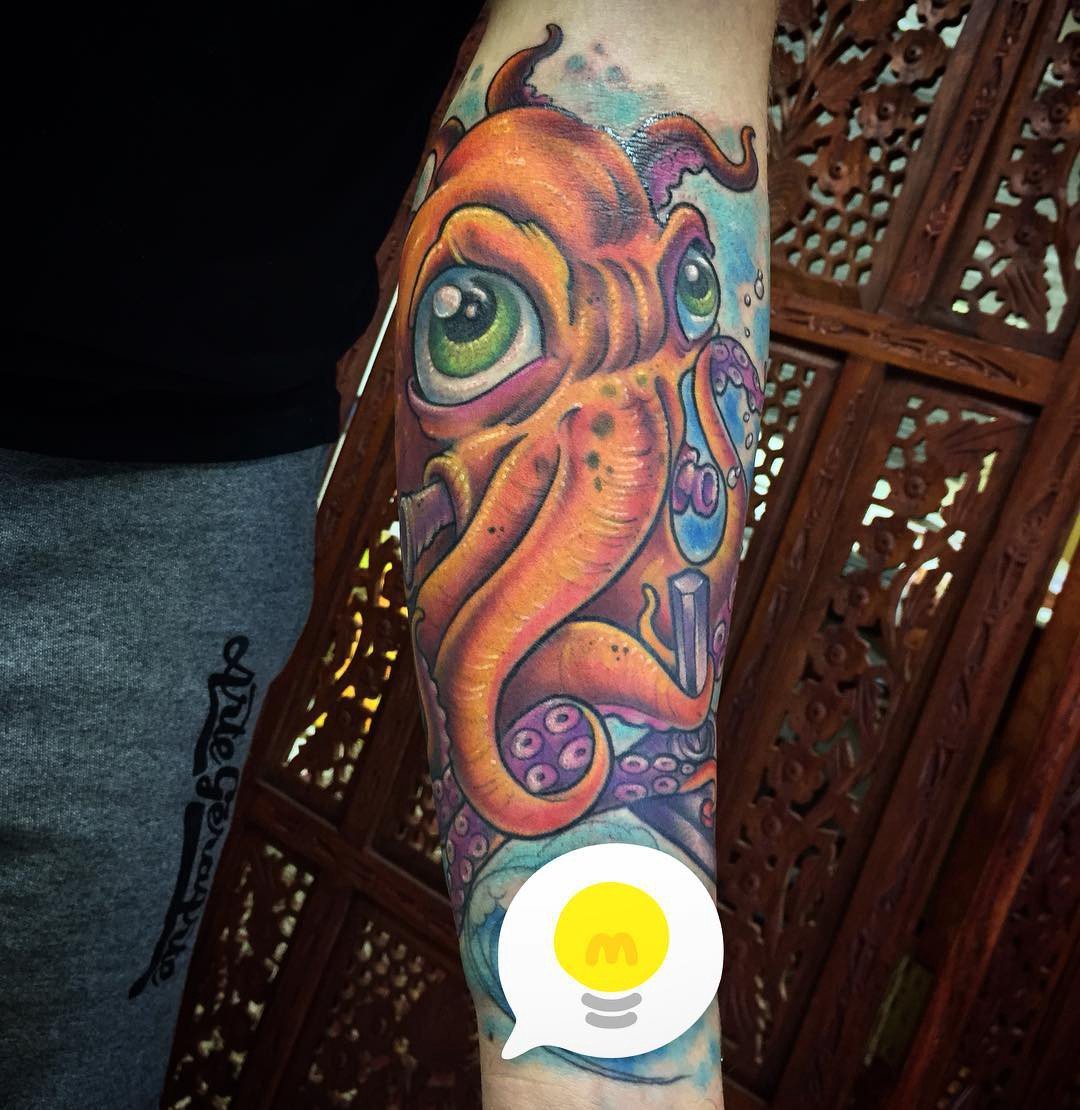 1gmmtzry0ovjirtdfudxf3q-1 Um braço tatuado com um polvo colorido e estilizado, visto sobre um fundo decorativo de madeira.