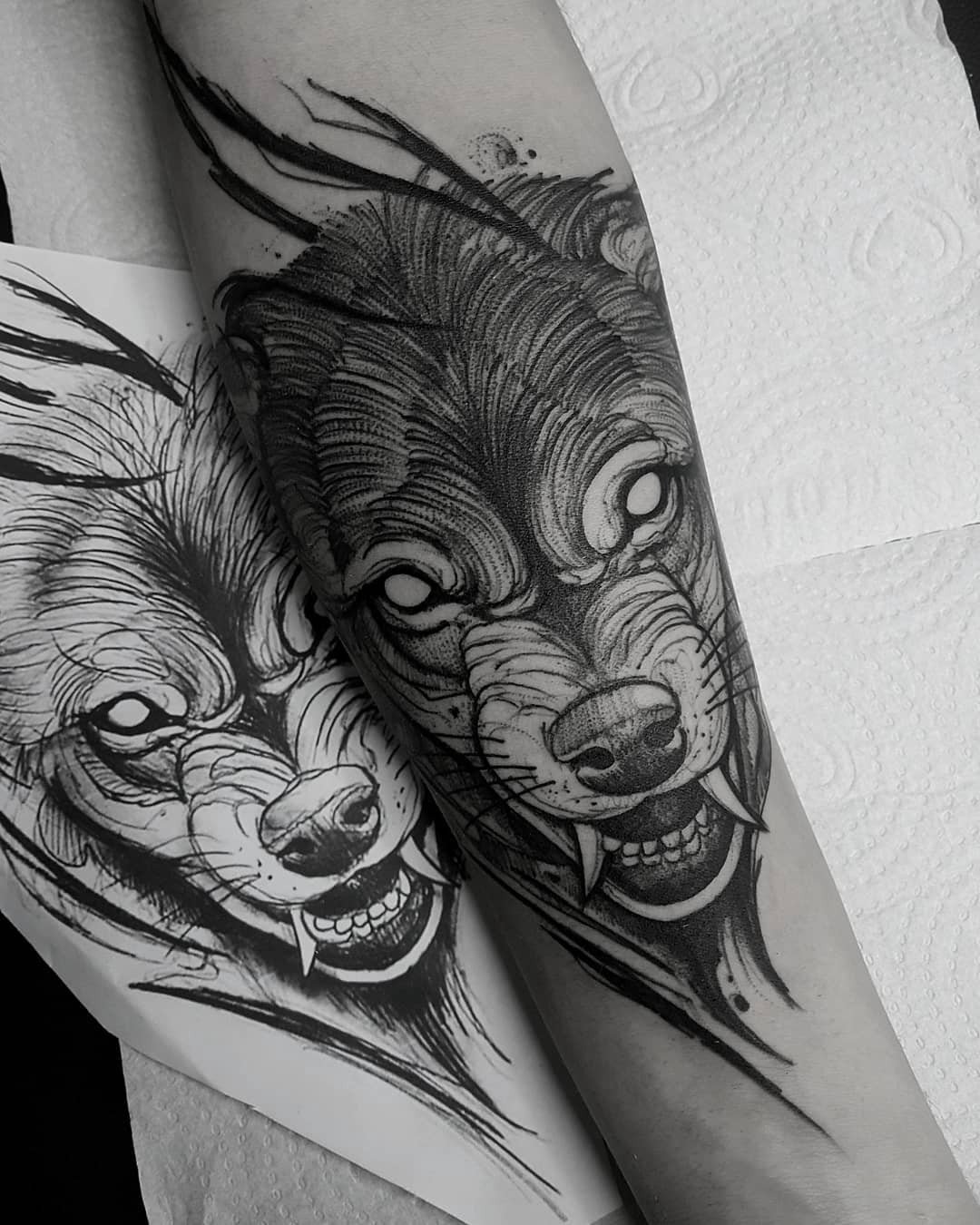Um braço com uma tatuagem de lobo detalhada ao lado de um desenho similar sobre papel.