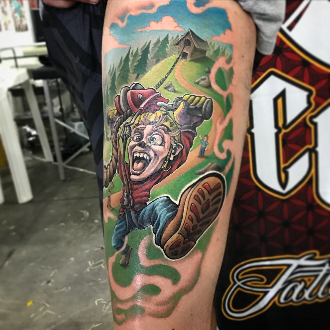 1k-czcsa2owsrttnhguvgvw-1 Uma tatuagem colorida mostrando uma figura animada segurando uma serra elétrica, com paisagem ao fundo.