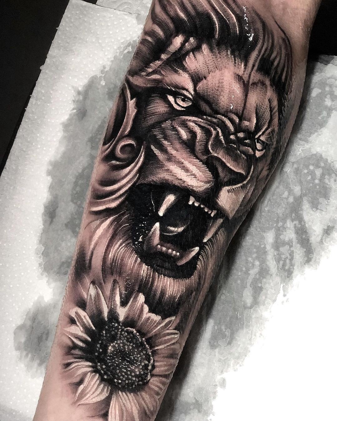 1lgidgyit9fmqp3vnzywvng-1 Tatuagem de leão rugindo com um girassol abaixo, detalhada em preto e cinza, no braço de uma pessoa.