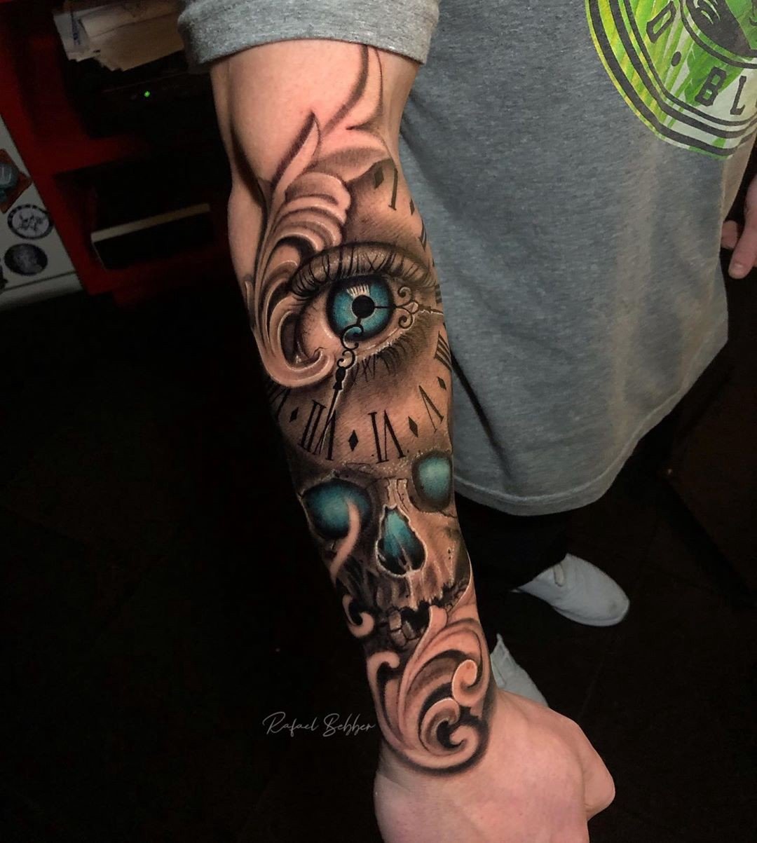 1lkpkcjkbmuowpqo7igqb0w Braço tatuado com desenho de relógio, olho azul e caveira em estilo realista e detalhado.