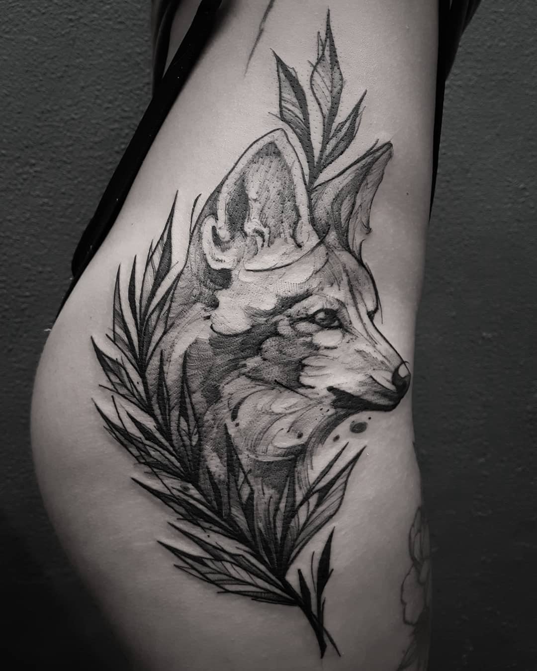 Tatuagem de lobo no corpo, com folhas decorativas ao redor, em estilo preto e cinza.