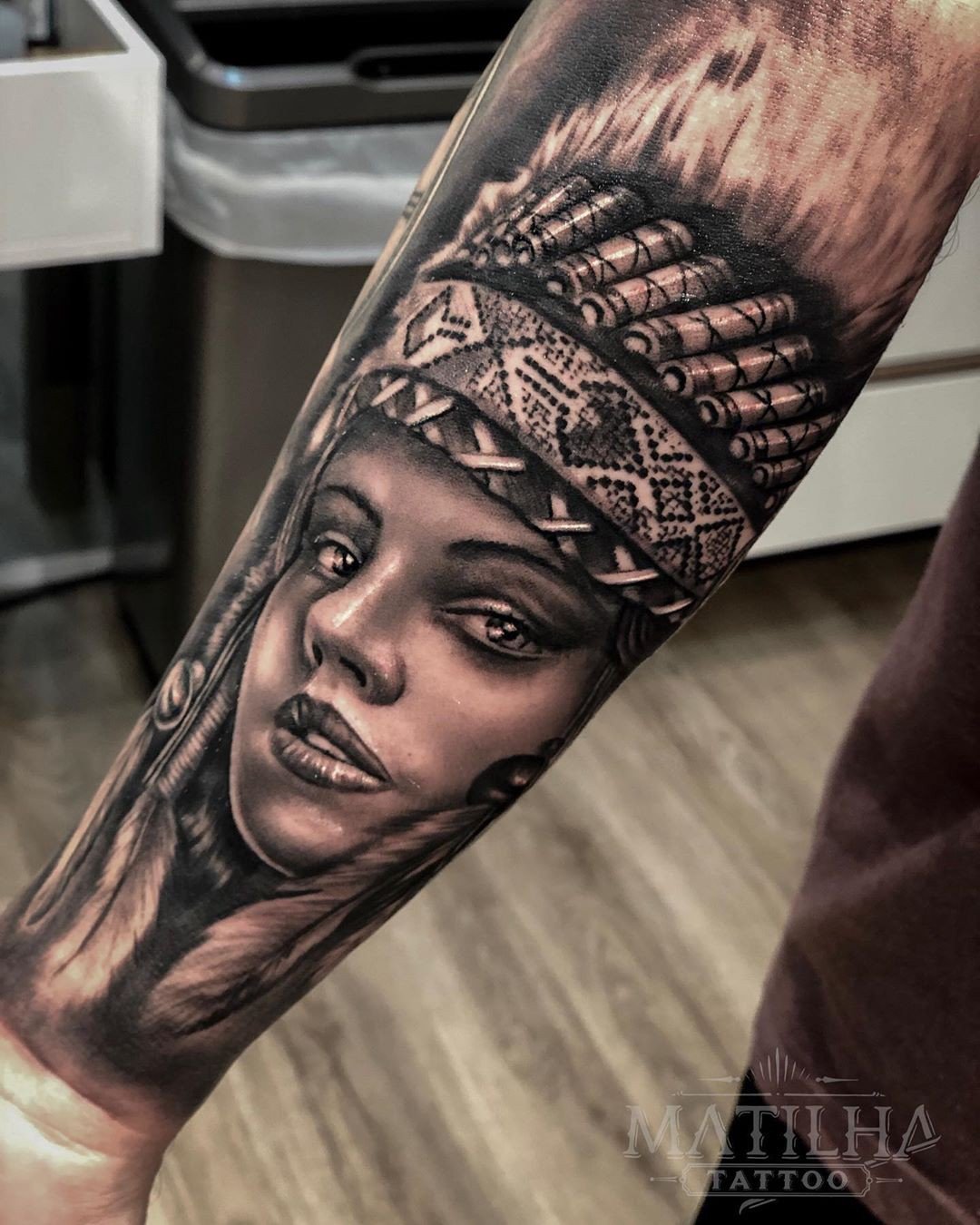 1n9mqj6js8a88n2ahbgherq Braço com tatuagem detalhada de rosto feminino com cocar, em estilo realista, fundo desfocado.