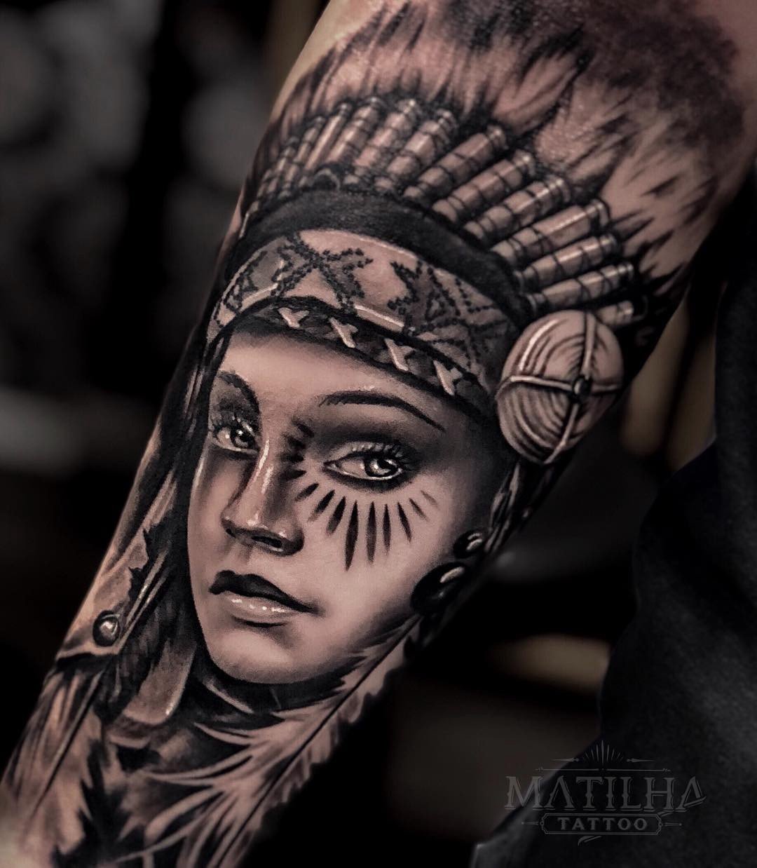 1nskgennuzqxbbxyovnzciw Uma tatuagem detalhada de um rosto com adereços de penas e pinturas faciais em preto e cinza.