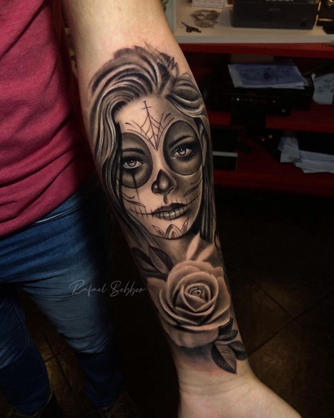 1ocpcxkx7snt2_mi-kyo1rg Braço com tatuagem de rosto estilizado com caveira e rosa detalhada, em tons de preto e cinza.