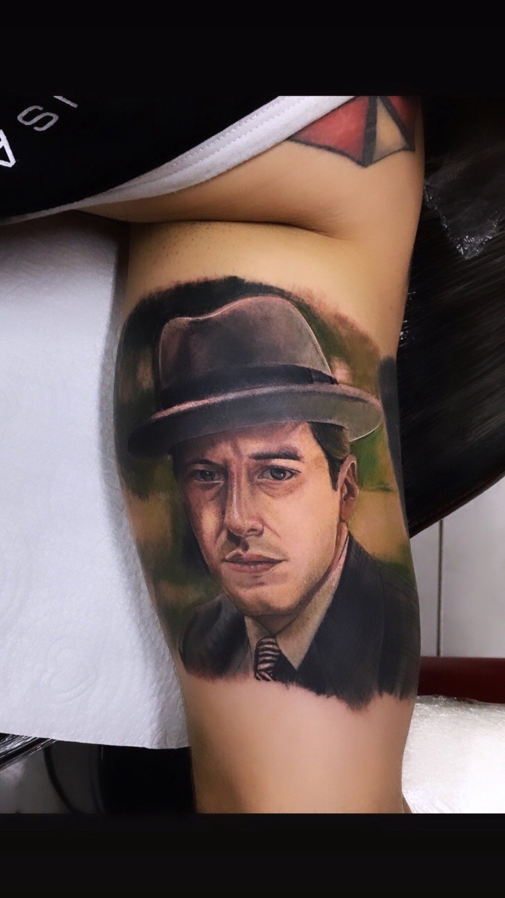 1papadfhatoyy1sndmggddg-1 Uma tatuagem no braço retrata um homem usando chapéu e terno, com expressão séria.