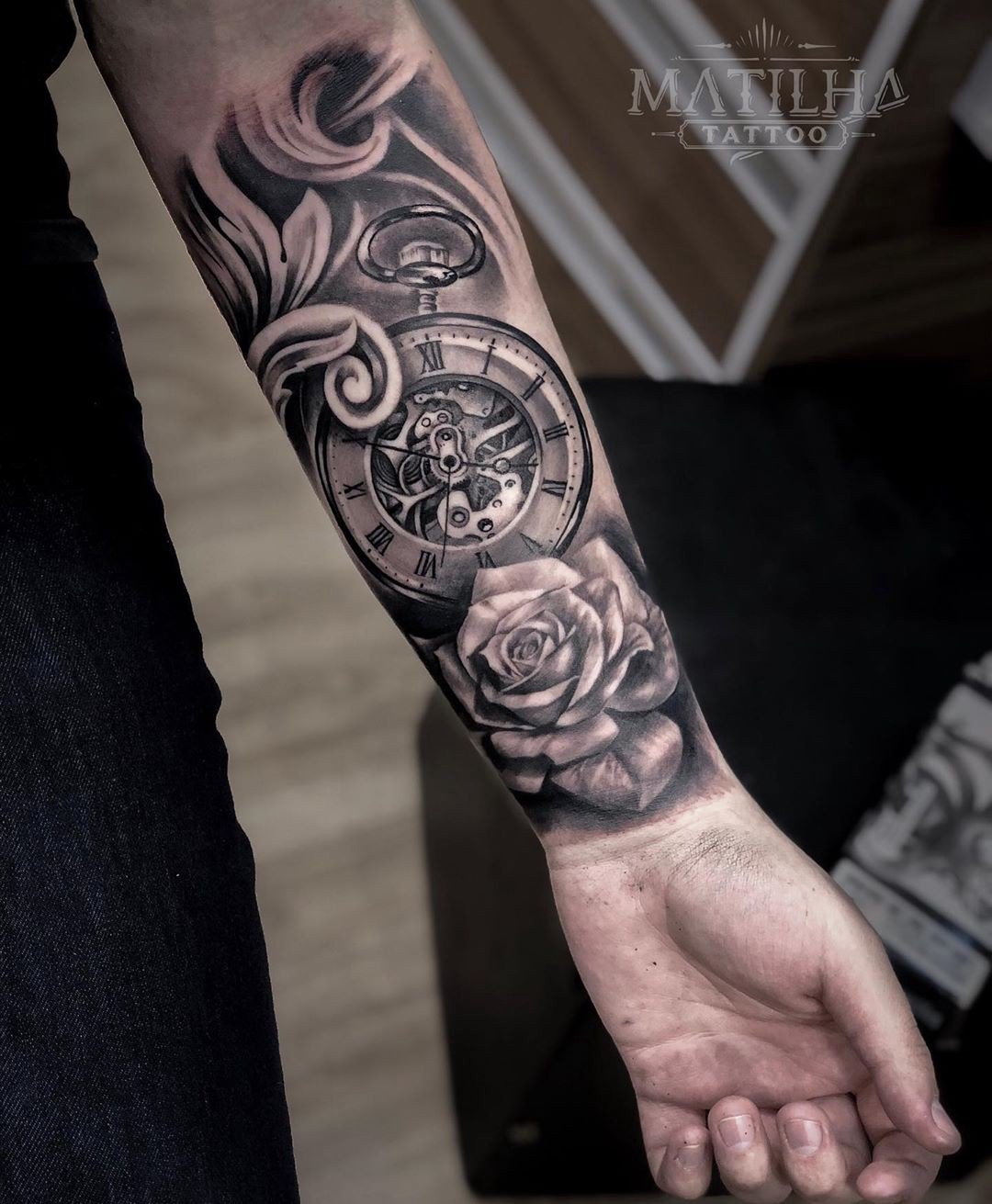 1qbb5mb6apxewsnd1koj7pw Braço tatuado com relógio de bolso e rosa em estilo blackwork, detalhes florais ao fundo.
