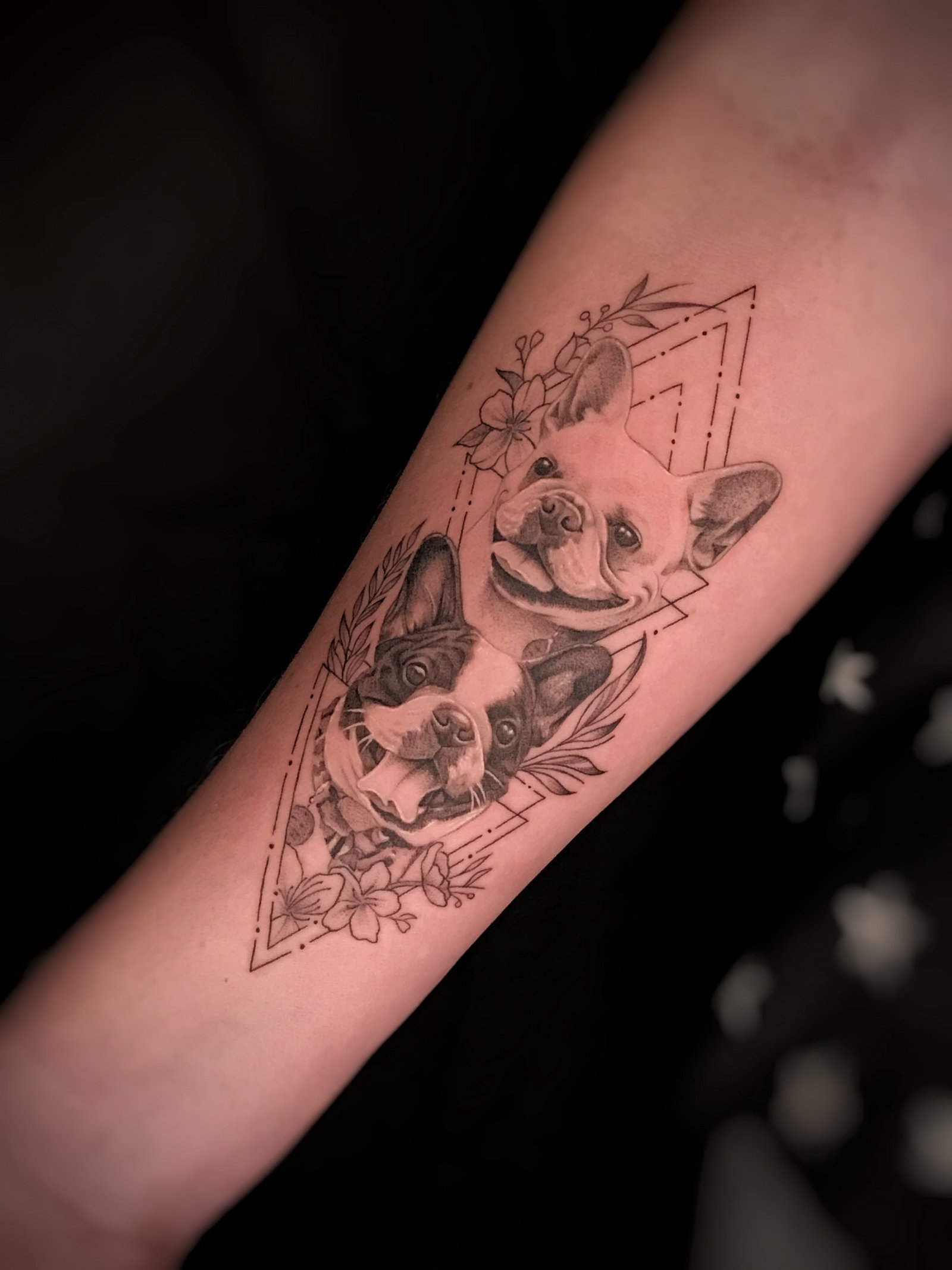 1qy81hke2hklfuej63gnp-q-1 Desenho de tatuagem no braço com dois cães cercados por flores e formas geométricas.