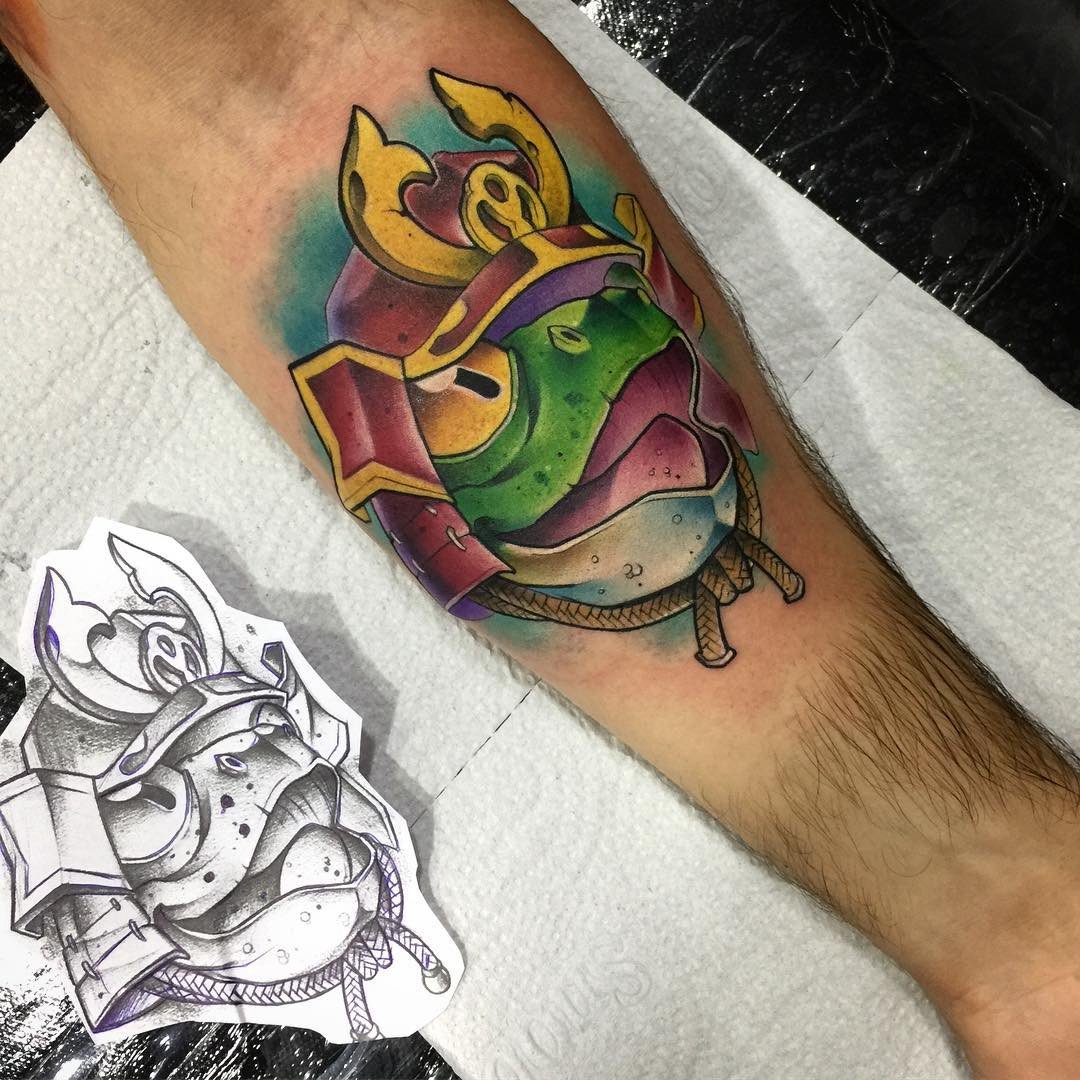 1rgikigujhbjefp__zgsr8a-1 Tatuagem colorida de sapo samurai no braço, com esboço em preto e branco ao lado.