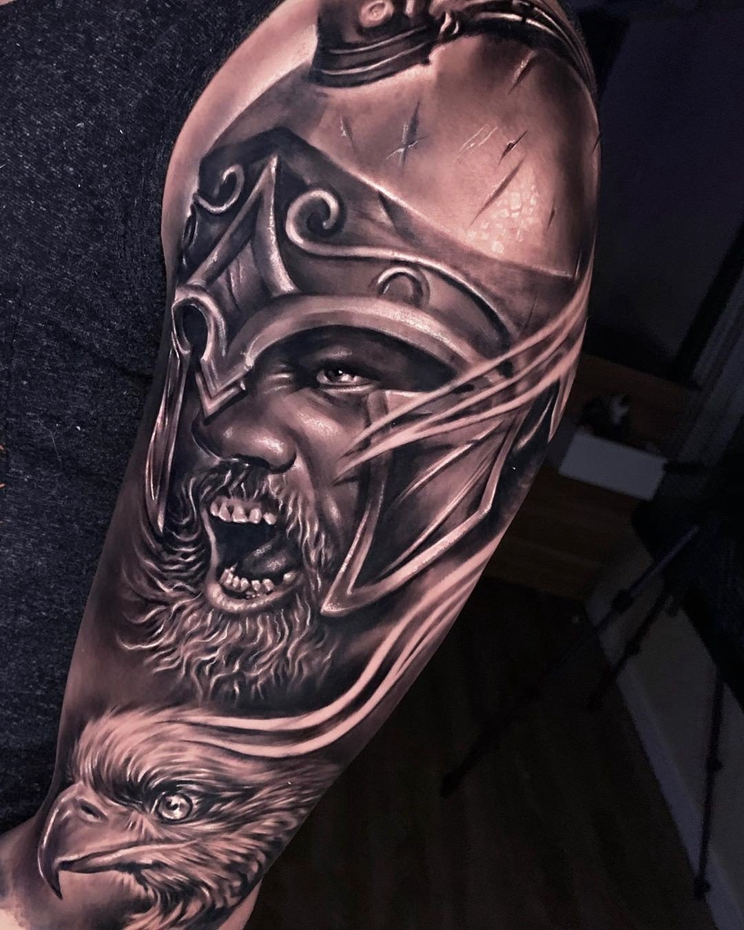 1sbvlxfuucpj2qvurzkfleq Tatuagem em braço de guerreiro com elmo e expressão de bravura, acompanhado por ave na parte inferior.