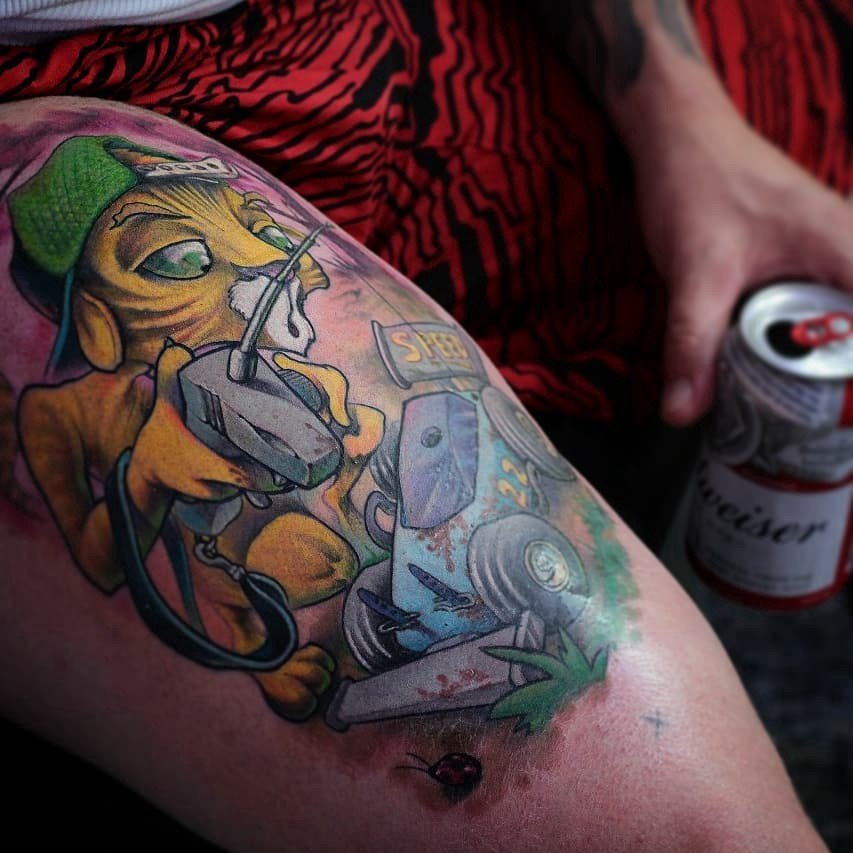 1sdxmnkewogtnbsk1idggoq-1 Tatuagem colorida de personagem animal com boné, segurando controle remoto e próximo a um carrinho de brinquedo.