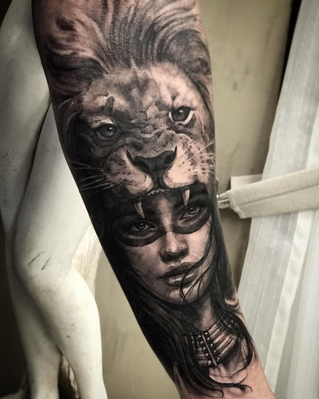 1svxmkyoa9nnbt7vku1ot7a Tatuagem de leão sobreposta ao rosto de uma mulher em um braço, com detalhes em preto e cinza.