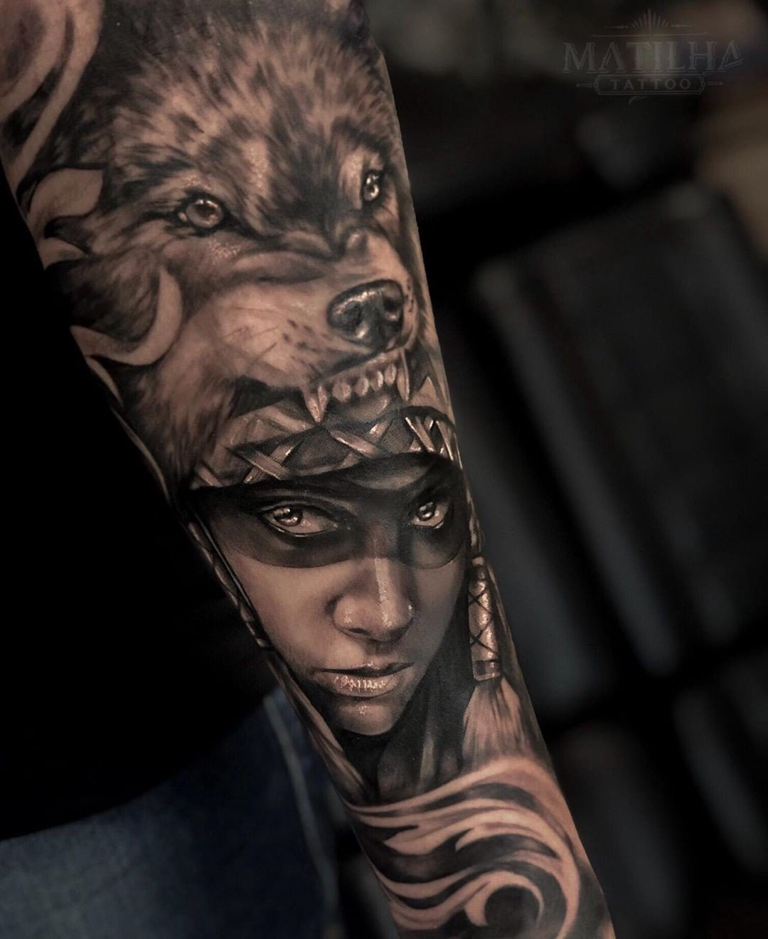 1t7y4edykifq-loocpp7zja Tatuagem em um braço com desenho de um lobo e rosto de pessoa usando adereço na cabeça.