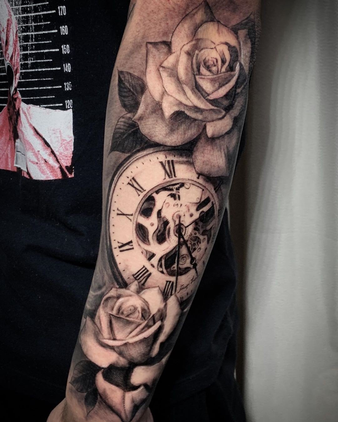 1tackl-qrme452dxs01t3za Braço com tatuagem de rosas e relógio com algarismos romanos, destacando detalhes sombreados em preto e cinza.