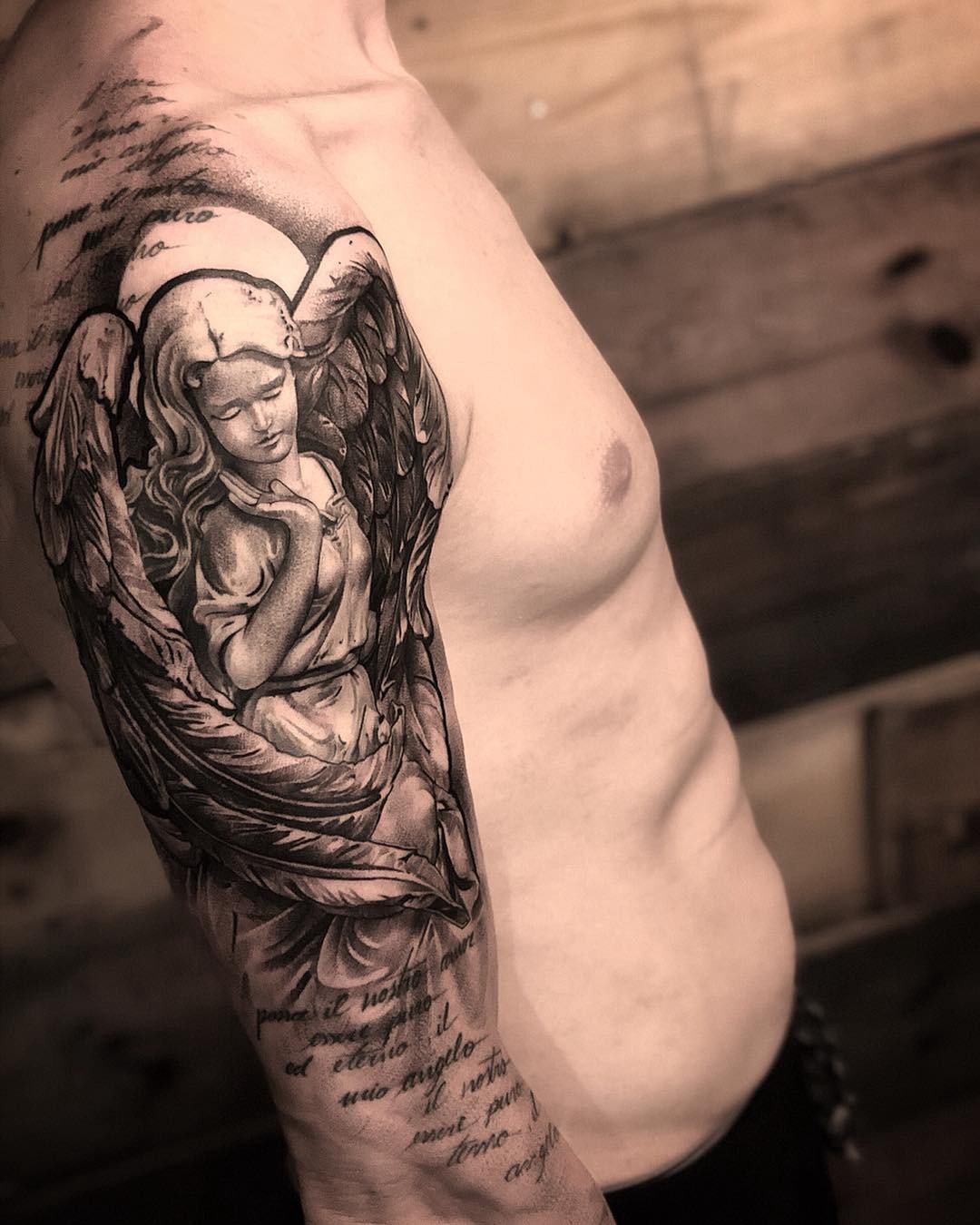 1tcuje-e3i_gcsbeelol2vq Tatuagem de anjo com asas e detalhes em sombreamento no braço de uma pessoa, acompanhada de texto.
