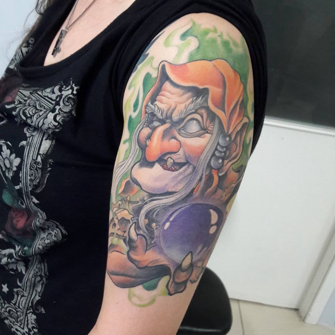 1ubqcbapvpj5owe1mo4enea-1 Tatuagem colorida de personagem com capuz, nariz proeminente e bola de cristal, no braço de uma pessoa.