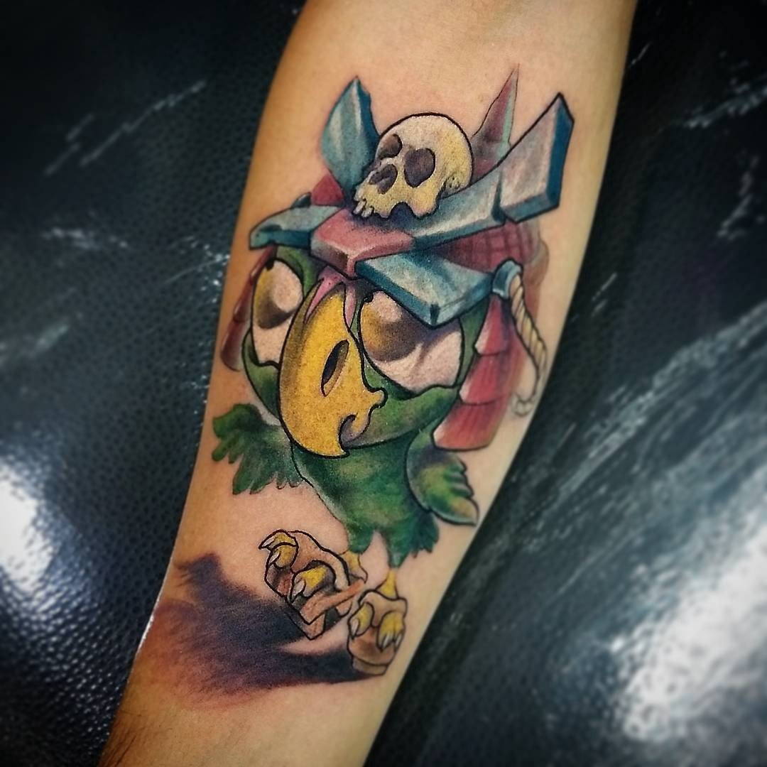 1ueicxr6x5-upfct89p3nhw-1 Tatuagem colorida de pássaro verde com capacete estilizado e caveira no topo, localizada no antebraço.