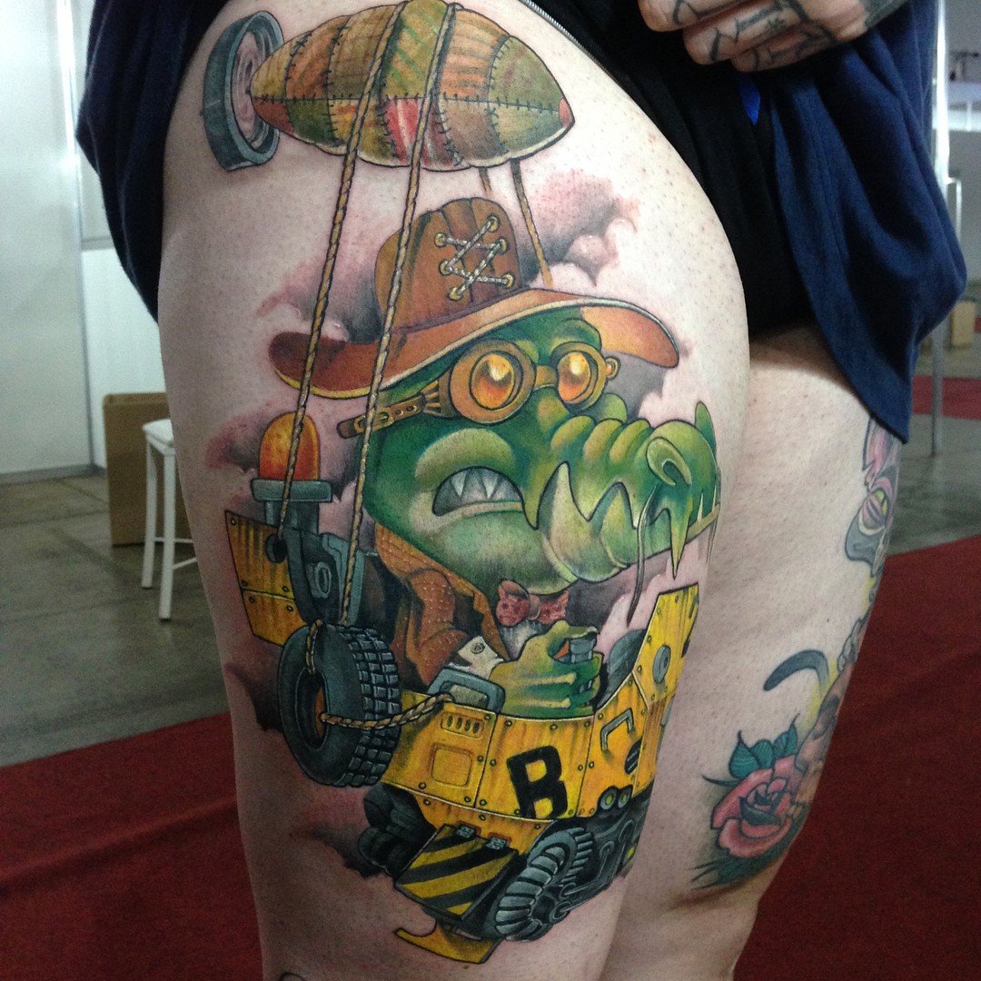 1yaqlxsi-ehk5-gb6ucxb_q-1 Tatuagem colorida de crocodilo antropomórfico em uma máquina voadora, com chapéu e óculos, na coxa de uma pessoa.