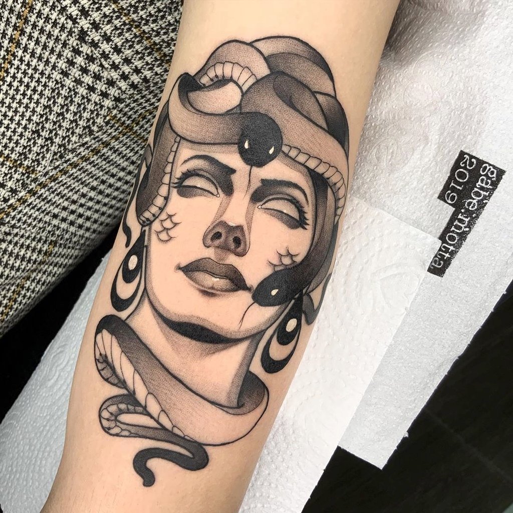 w1024-141 Tatuagem no braço de uma figura feminina com serpentes ao redor da cabeça, em estilo preto e cinza.