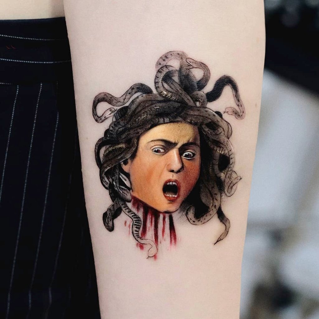 w1024-158 Tatuagem no braço de cabeça com cobras no cabelo e expressão de susto, com detalhes em vermelho na base.