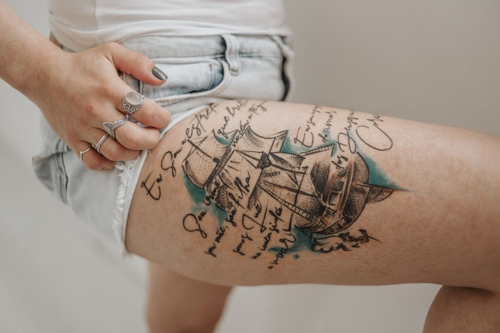 Tatuagem na coxa de um navio com inscrições em torno, shorts curtos e pessoa com anéis prateados na mão.