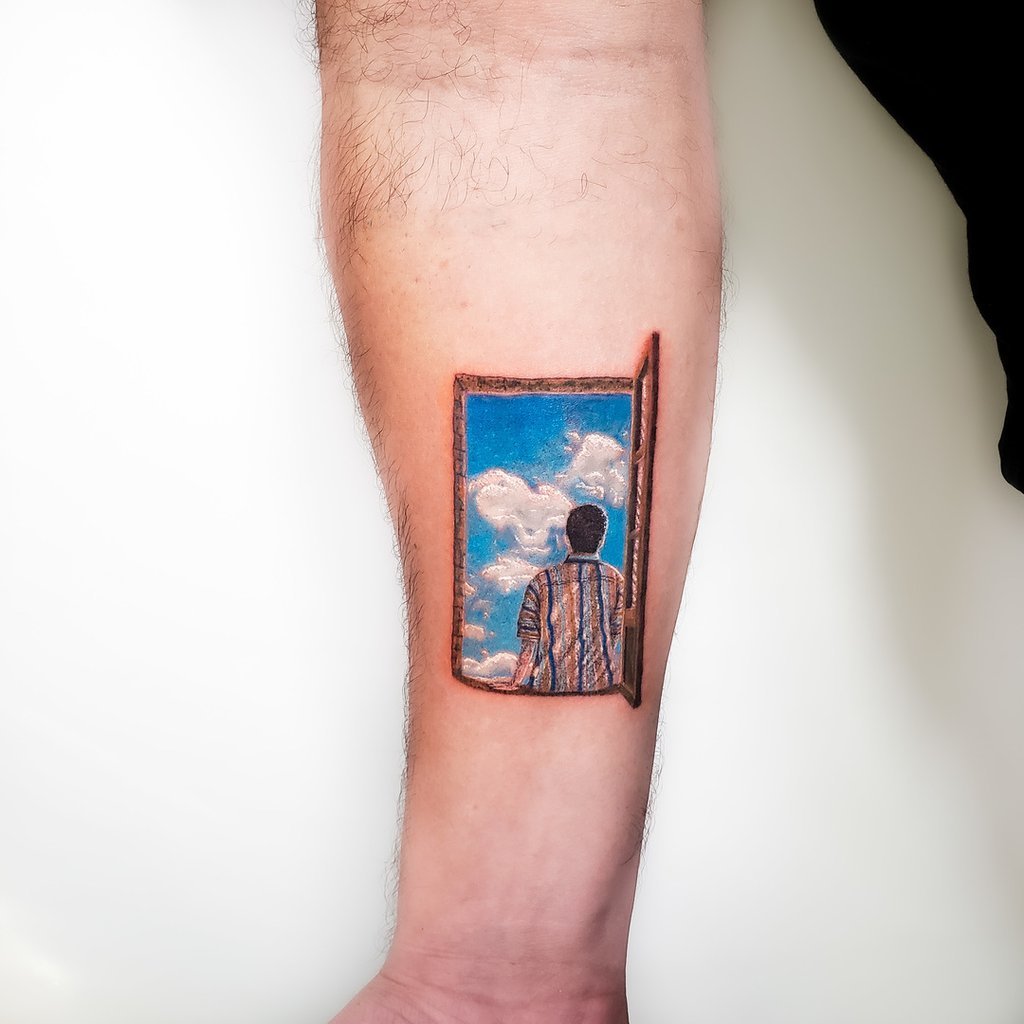 w1024-30 Uma tatuagem no braço mostra uma pessoa de costas olhando para o céu azul com nuvens através de uma janela.