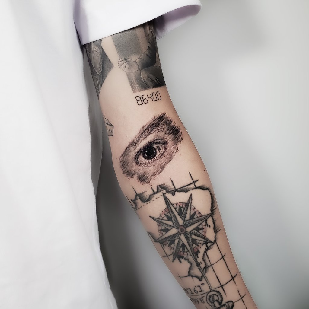 w1024-33 Um braço tatuado com um olho, uma bússola e números, em frente a um fundo branco.