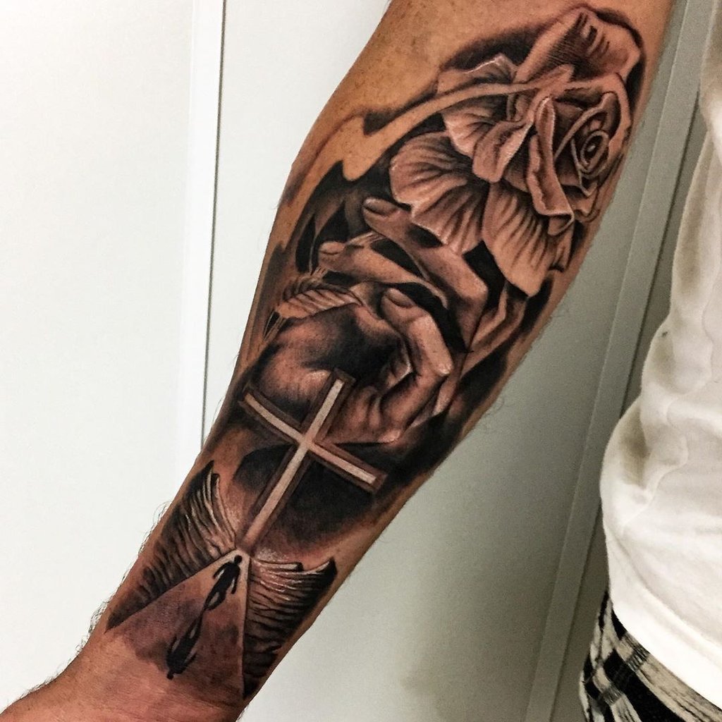 w1024-54 Um braço tatuado com uma rosa, mãos em oração, uma cruz e asas, formando uma composição artística detalhada.