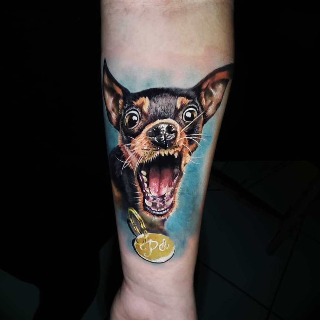 w1024-55 Tatuagem colorida de um cachorro de boca aberta com uma medalha dourada, localizada no antebraço de uma pessoa.