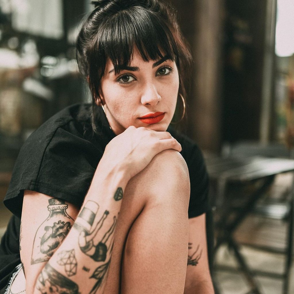 w1024 Pessoa com tatuagens, cabelo escuro e batom vermelho, olhando para a câmera em ambiente interno.