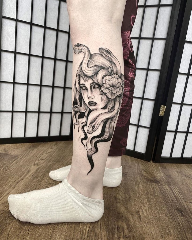 w819-112 Perna com tatuagem de figura mitológica, serpentes e flor, em fundo de divisórias e chão de madeira.