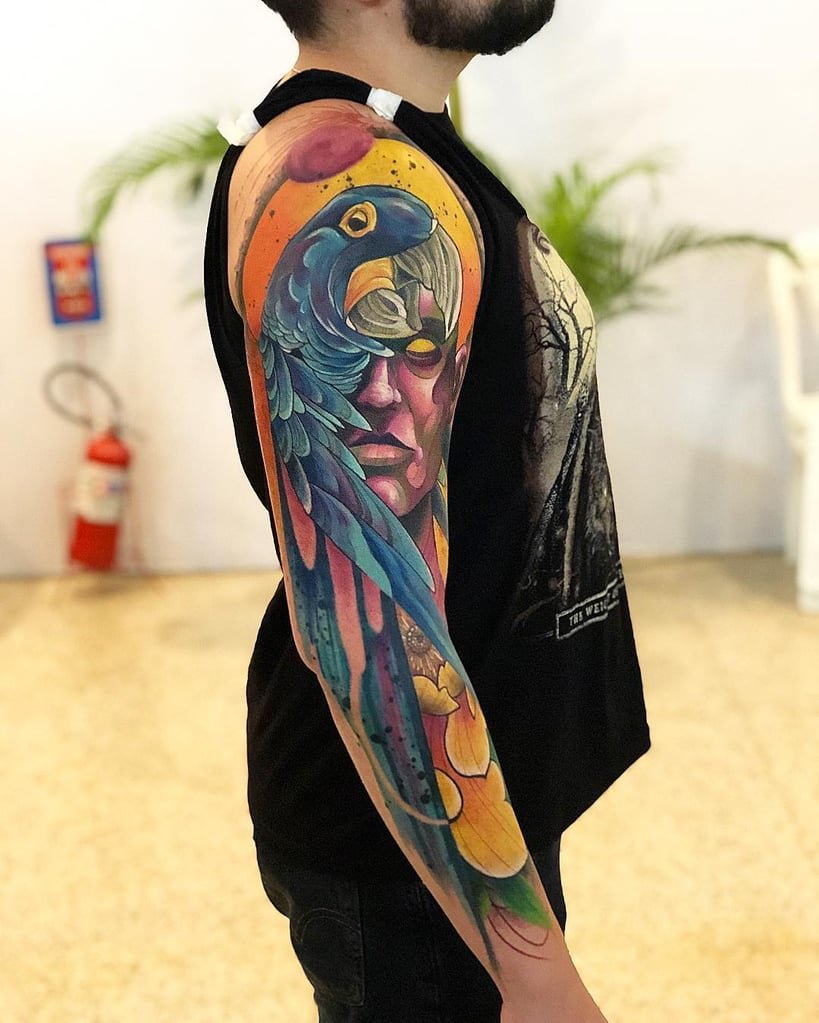 w819-16 Pessoa com tatuagem colorida de papagaio e rosto humano no braço, em ambiente interno.