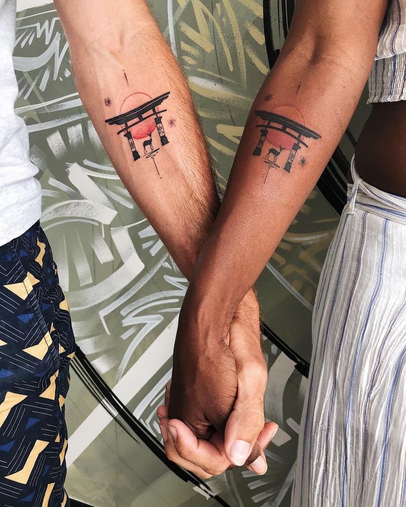 w819-208 Duas pessoas de mãos dadas, cada uma com uma tatuagem de torii no antebraço, em frente a um fundo artístico.