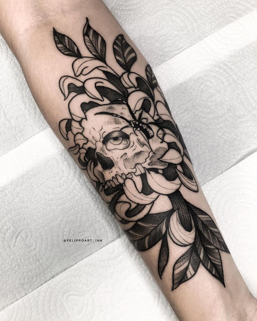 w819-24 Tatuagem de caveira com folhas e flores em estilo preto e cinza no braço de uma pessoa.