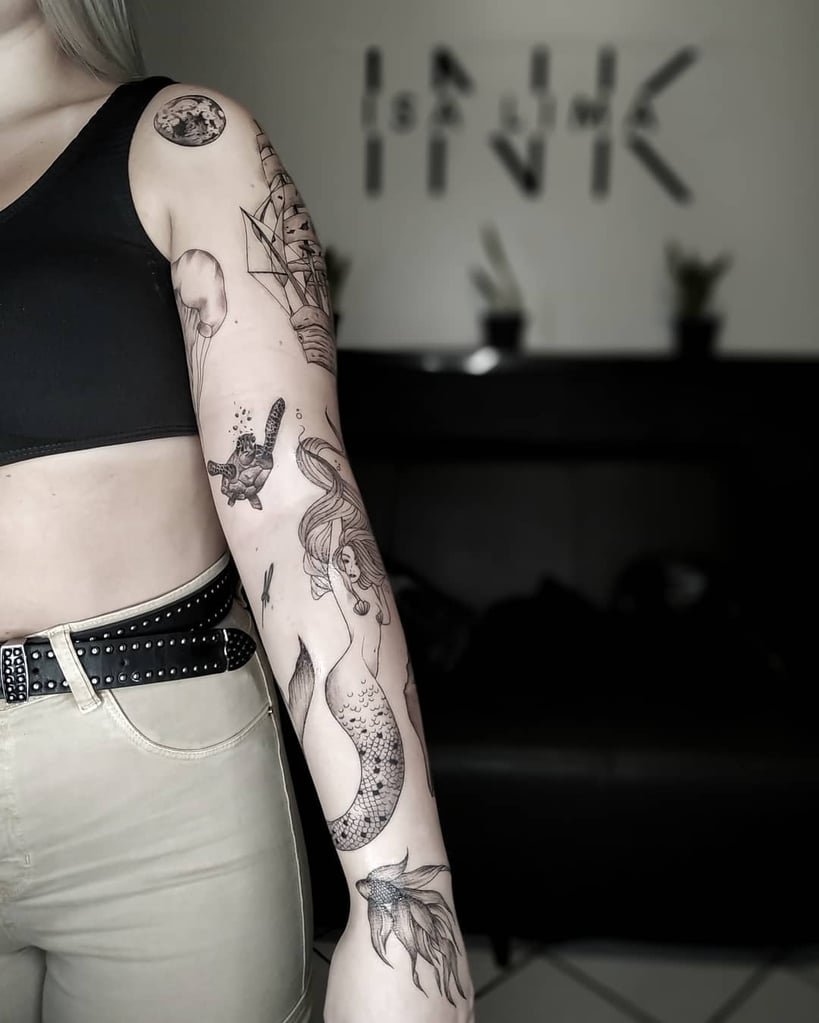 w819-33 Pessoa com várias tatuagens detalhadas no braço, incluindo sereia e navio, em ambiente interno.